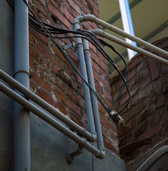 Canalizaciones sanitarias en fachada de ladrillo visto con cableado desprotegido, mostrando una instalación precaria sometida al rigor atmosférico. Multiservice A.D. advierte la necesidad imperiosa de inspeccionar redes expuestas.
