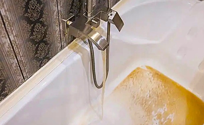 Caudal de agua con alta saturación ferrosa de tonalidad ámbar emanando de una grifería y manchando el fondo de una bañera doméstica.