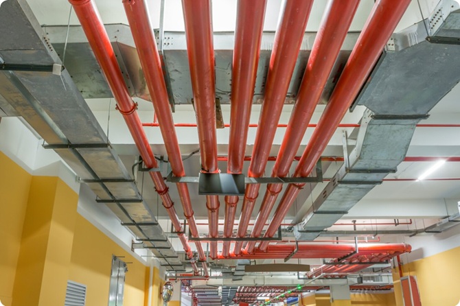 Red de protección contra incendios suspendida en el techo de un garaje industrial, con tuberías de acero en color rojo vibrante y conductos de ventilación.
